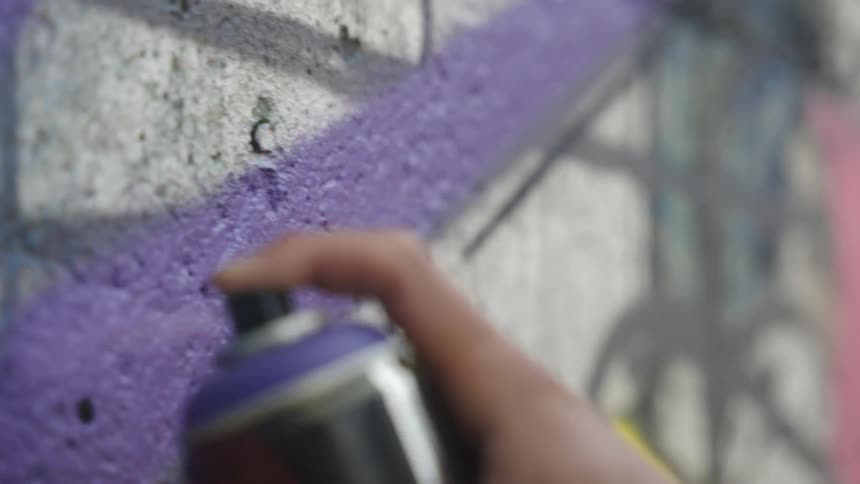 A hand using a can of purple paint to paint a graffiti on a wall