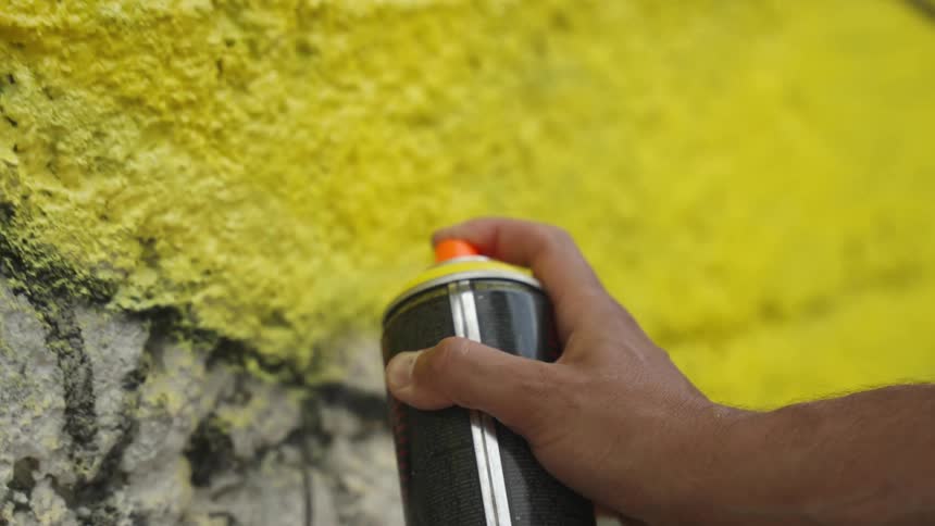 Close up of a mans hand with yellow spray paint graffitiing a wall
