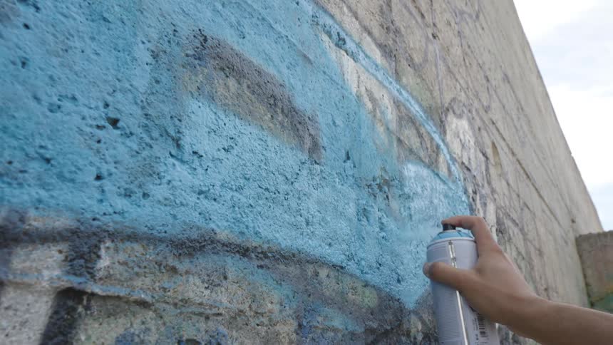 A hand using a can of blue paint to paint a graffiti on a wall