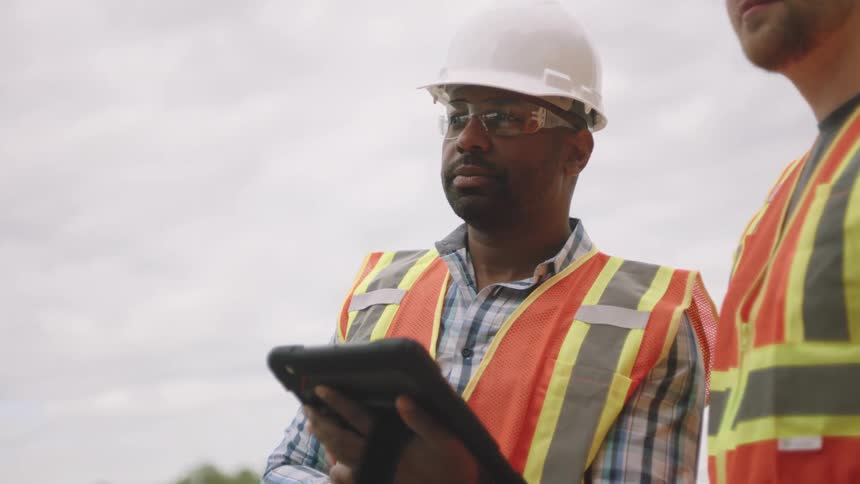 Two male workers dressed in vests and helmets with one of them holding a tablet