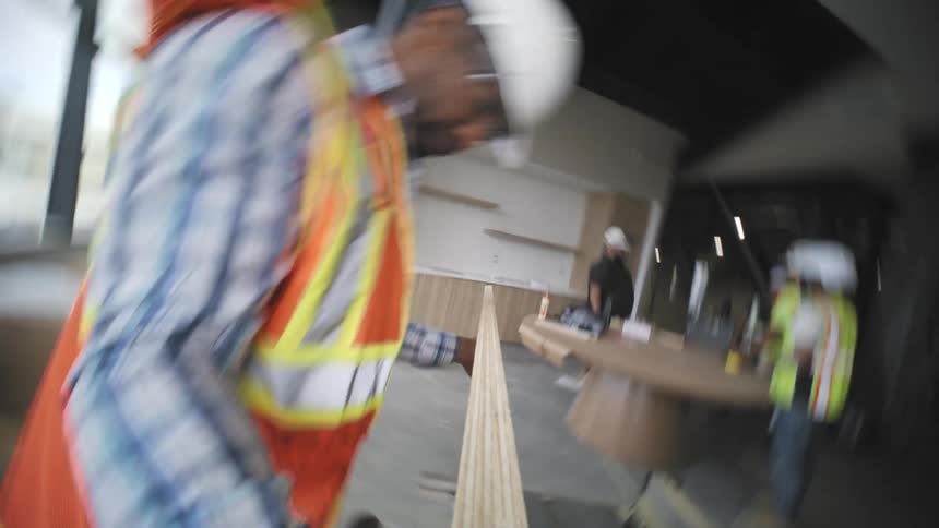 A person wearing a white hard hat and highvisibility vest is cutting and transporting wood at an indoor construction site