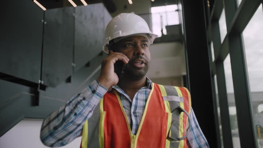 A construction worker wearing a helmet and a vest talks on the phone in a building