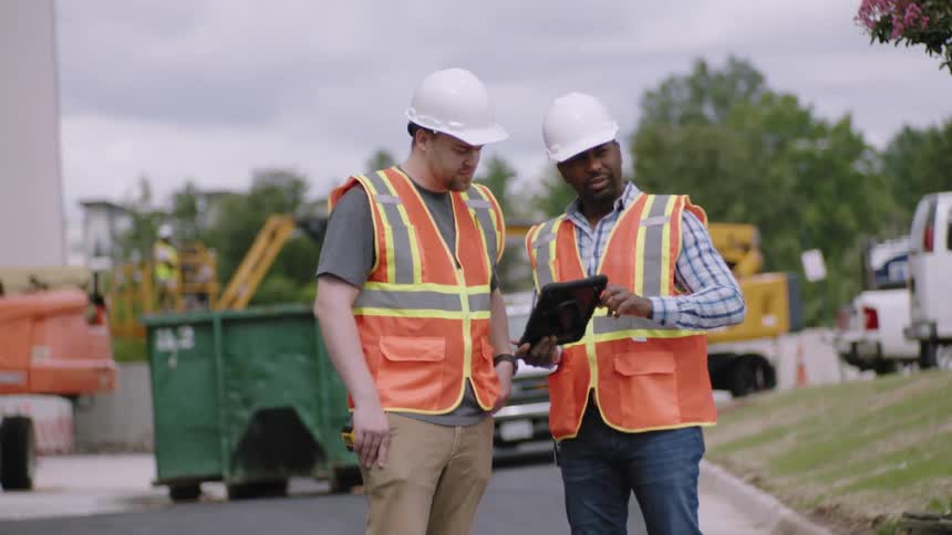 Two men one of them holding a tablet talking while both wearing a protective helmet