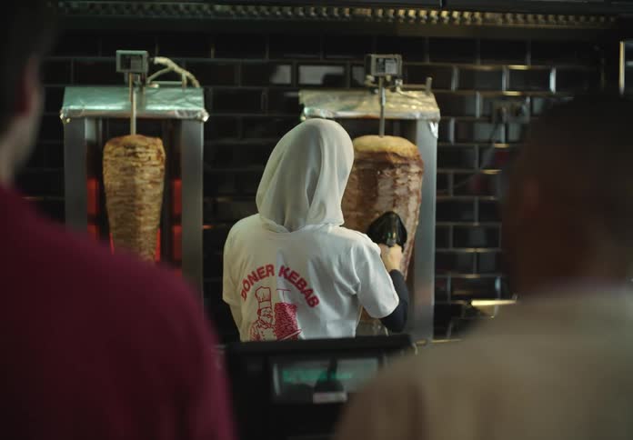 A woman wearing a white doner kebab shirt and a headscarf is preparing food in front of two rotating dner kebabs in a restaurant kitchen