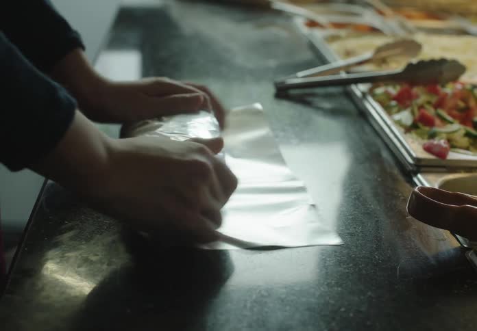 A closeup of hands wrapping a burrito or sandwich in aluminum foil on a restaurant counter