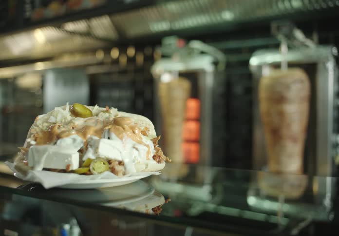 Plate full of food on a glass counter and in the background two pieces of meat being grilled in a restaurant