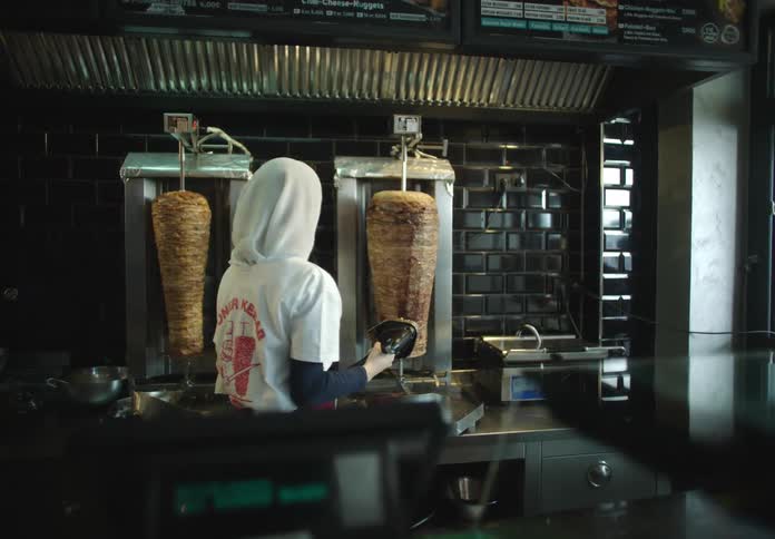 A woman in a white sweater with a hood stands in front of two large vertical spits of meat for kebab