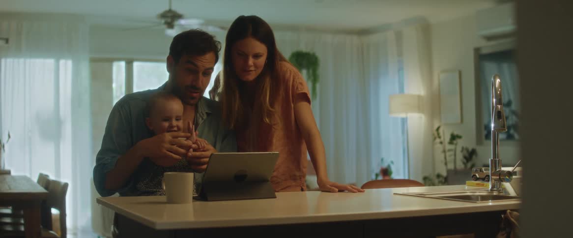 A father mother and daughter sitting and standing at their kitchen counter in front of a tablet smiling and laughing