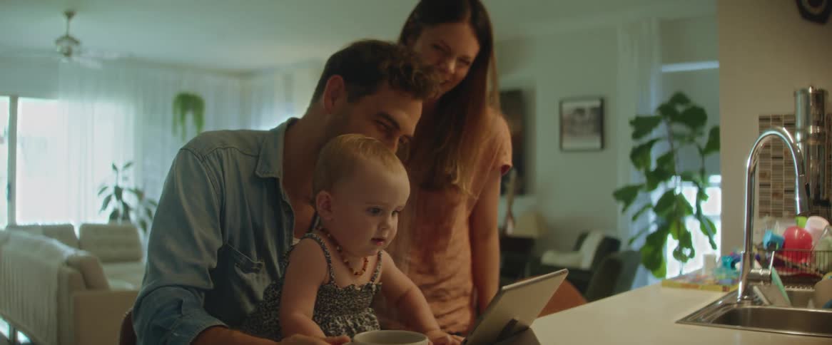 A father with his baby daughter on his lap and the mother next to them as they smile and laugh at the tablet on the counter