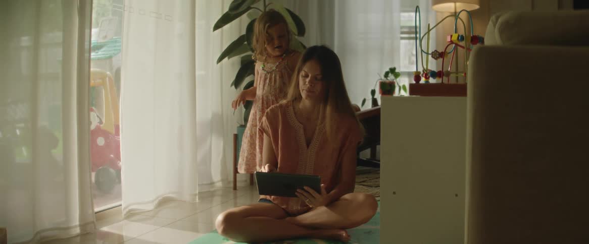 A mother sitting on the floor looking at her tablet while her young daughter plays with her hair