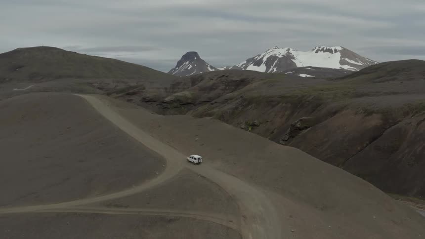An aerial of a white jeep driving through the desert on a road overlooking snow covered mountains