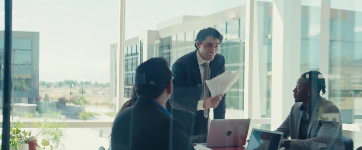 Man in a suit holding papers and talking cheerfully with other people sitting at a table