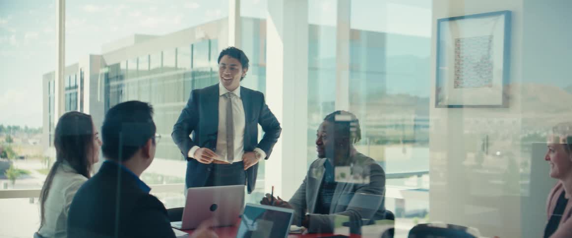 Man in a suit standing and talking to other people who are sitting at a round table in an office room