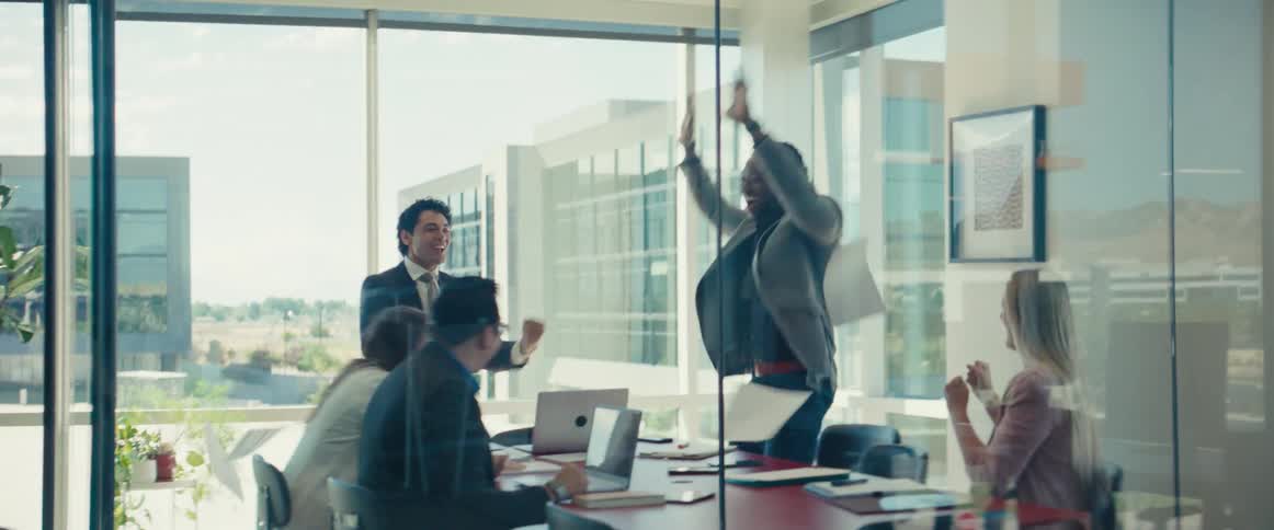 Man throwing papers in the air and people around him get up from the table and start celebrating in an office setting
