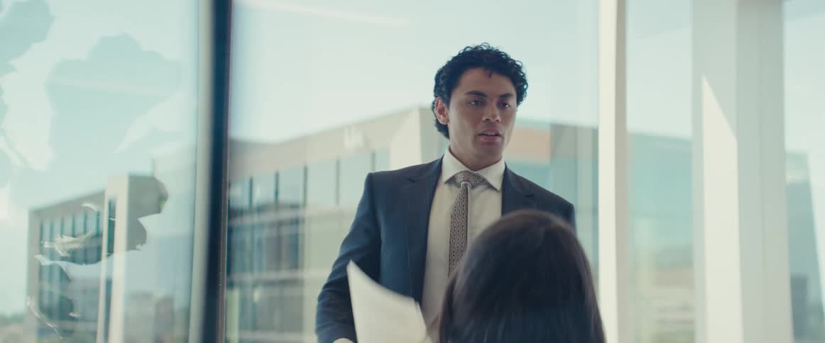 A man in a glass meeting room pacing side to side while holding papers and talking to other people