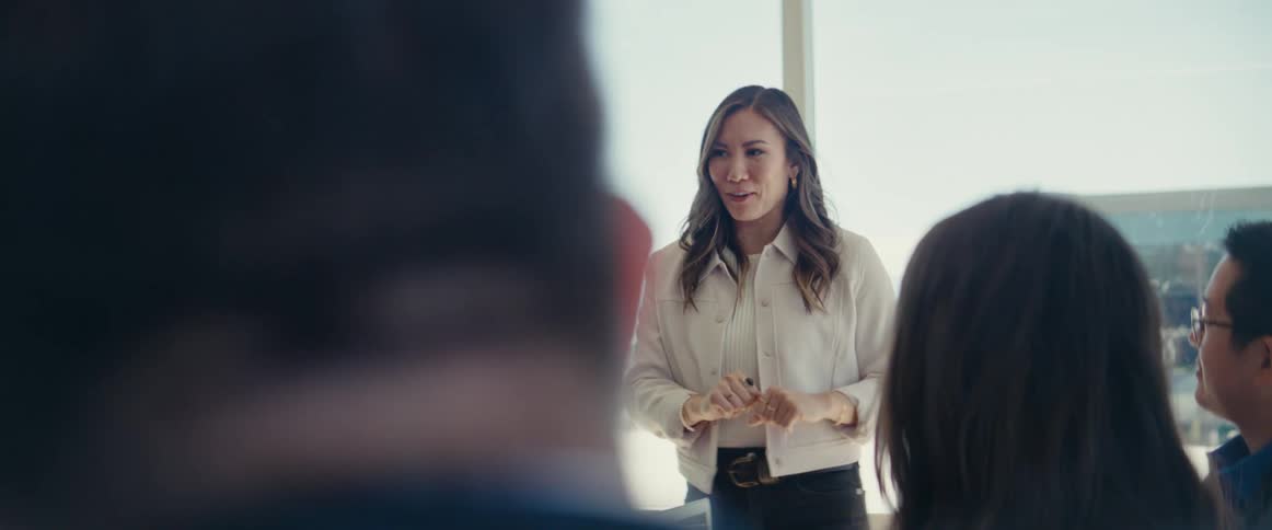 A business meeting in a glass office with one woman standing gesturing and smiling while talking to the others