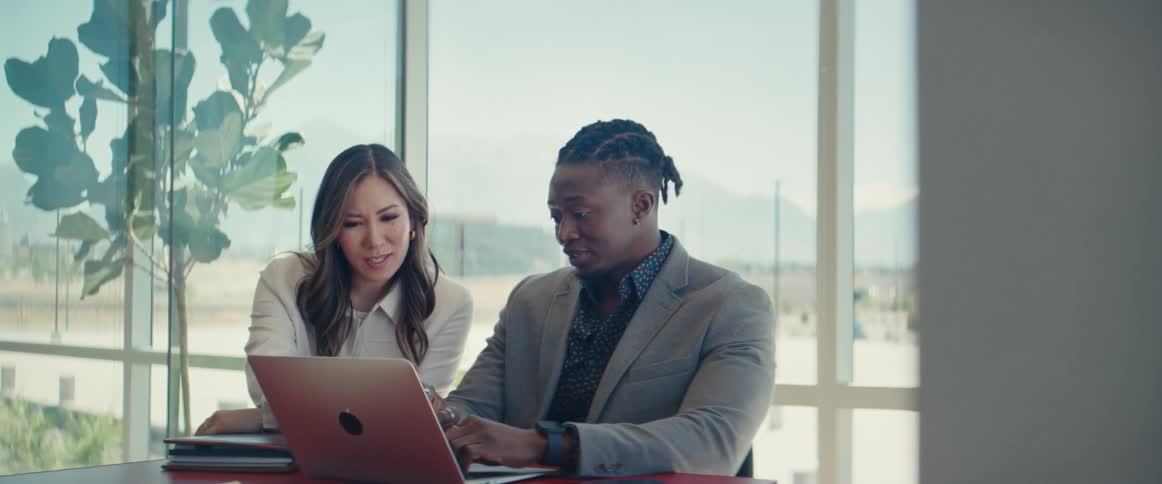 A woman and a man in a glass office sitting at a desk and discussing what they see in a laptop