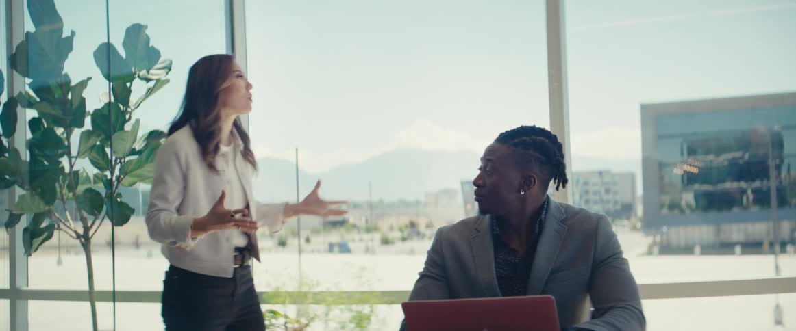 Woman walking back and forth talking to a man who is sitting in front of a laptop