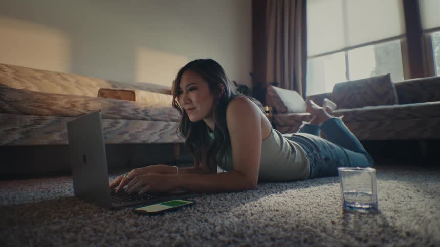 Woman lying face down on carpet typing on laptop with smartphone next to her in living room
