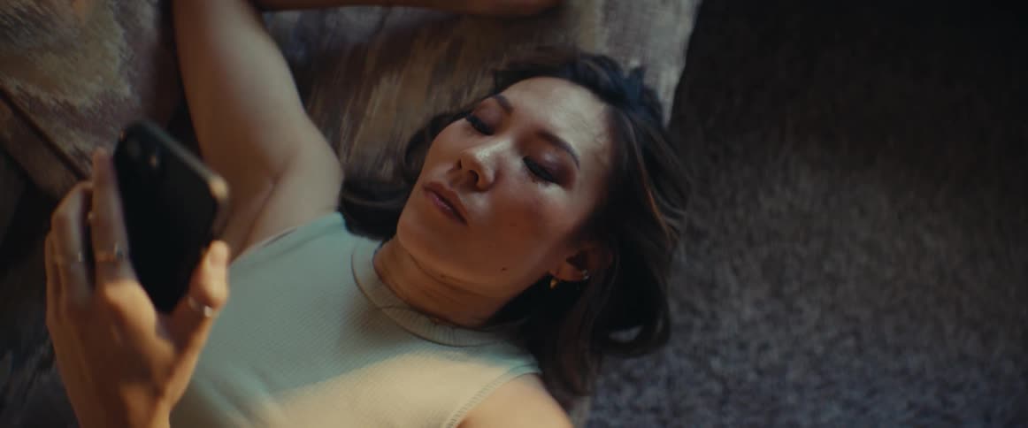A woman is laying on her couch thinking and eventually picks up her smartphone and types something