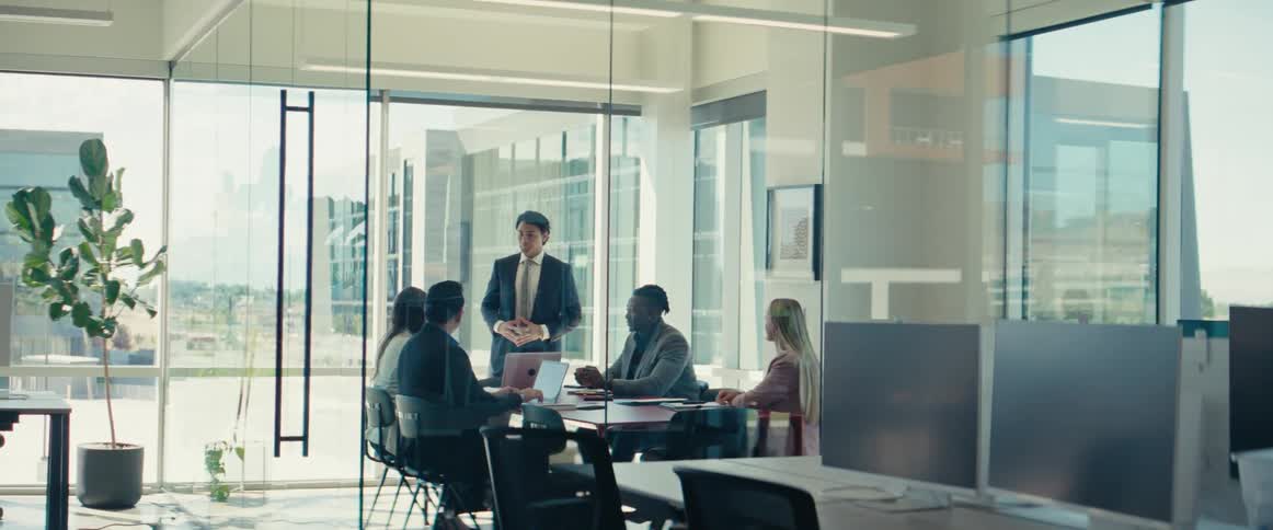 Man in a suit standing and talking to other people who are sitting at a meeting table in an office room
