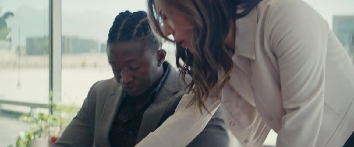 A woman assists a man in signing documents at a desk in a modern office setting