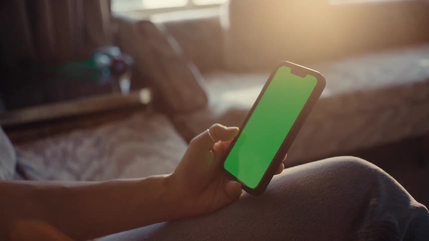 A person holds a smartphone with a green screen in an indoor setting with warm lighting