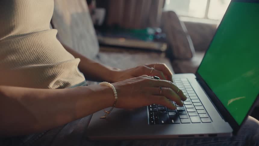 A woman sitting on a couch with a laptop on her lap typing and looking the replaceable green screen
