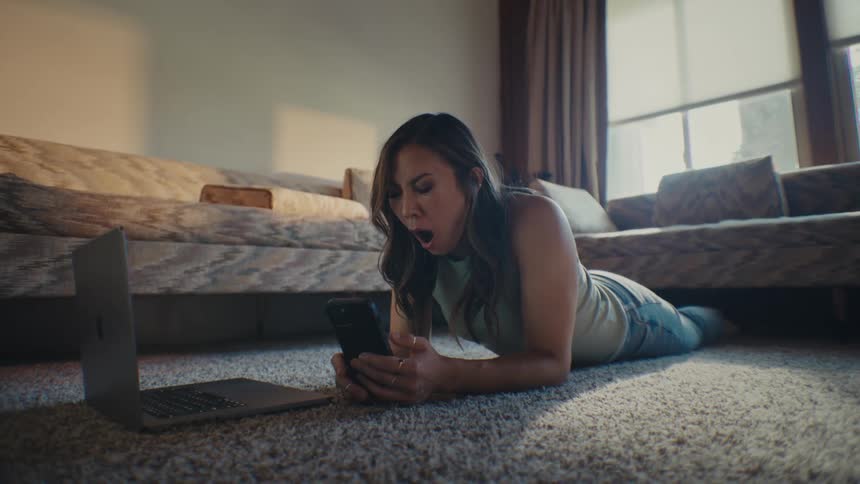 Woman lying on the living room carpet yawning and interacting with a smartphone on a laptop next to it