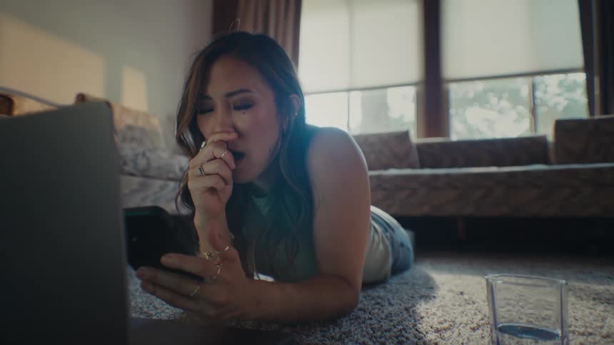 A young woman laying on her stomach in her living room while yawning looking at her cell phone and laptop in front of her