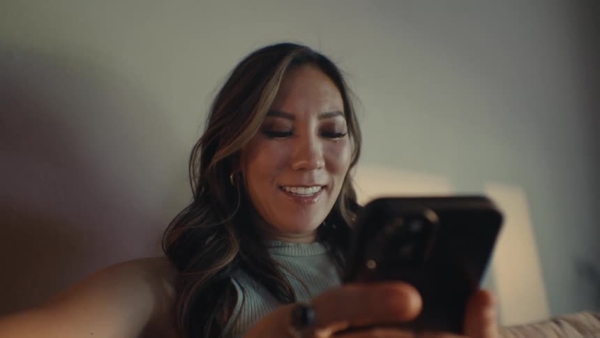 A young asian woman sits on her sofa while smiling and typing on her smartphone