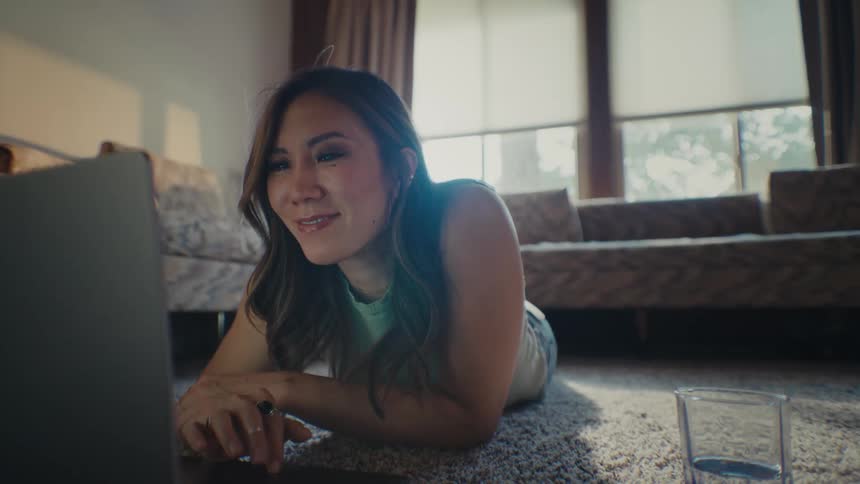 An asian woman lies on the living room floor while working on her laptop