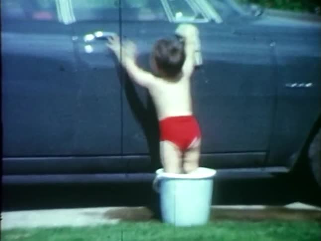 A toddler stands in a bucket to wash a car