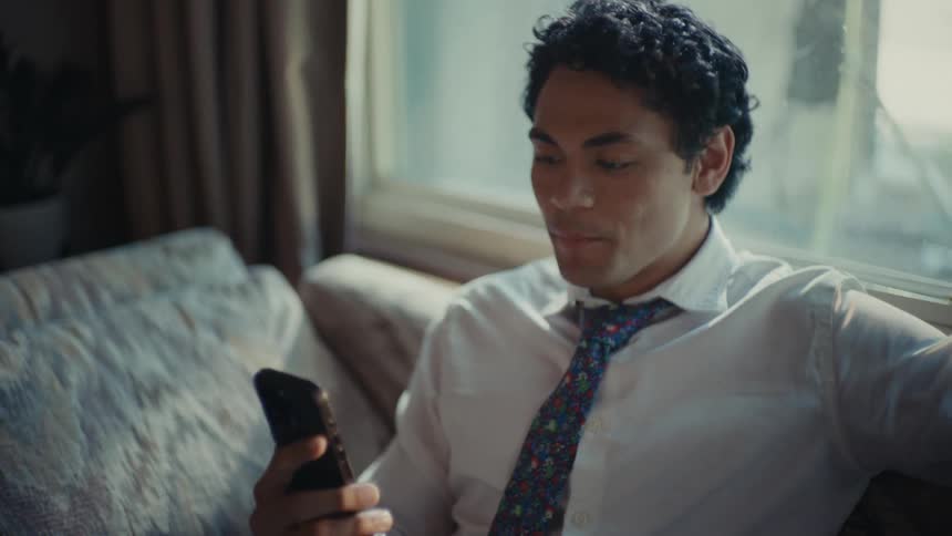 A hispanic man sits on the couch while talking on his smartphone