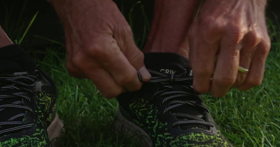 An elderly man sitting on grass and tying his shoelaces