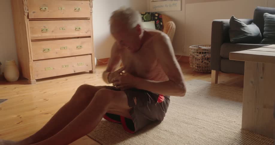 An elder man doing sit ups in his living room on a wooden floor surrounded by furniture