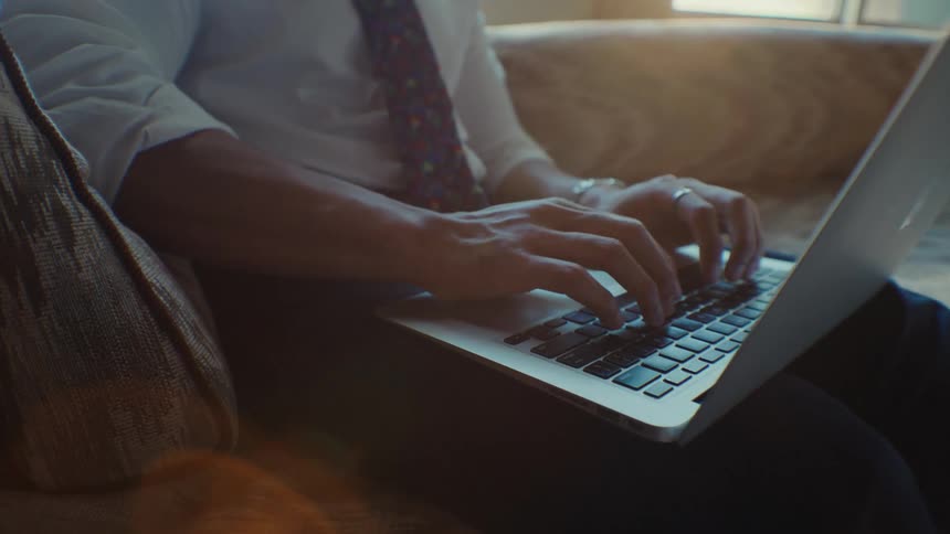 A man wearing a shirt and tie sits on a couch and types on the laptop on his lap