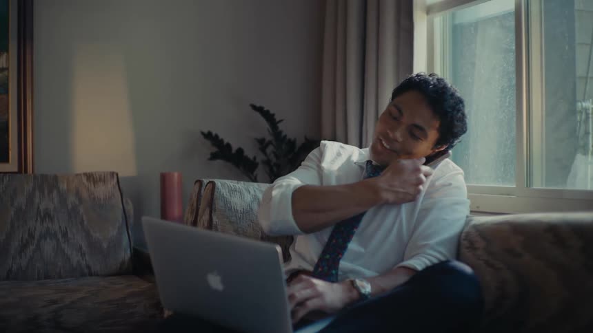 A man sitting on his couch talking on his cellphone while typing on his laptop