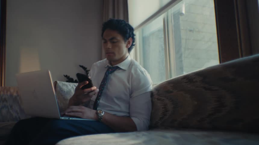 A man wearing a tie and slacks types on a laptop and calls someone on a smartphone