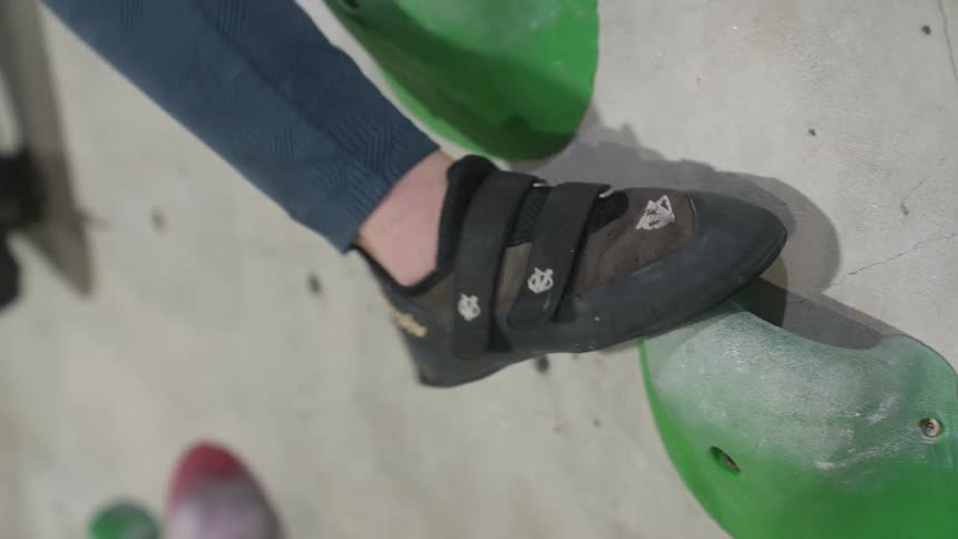 Closeup of a persons foot wearing a black climbing shoe with velcro straps stepping on a green climbing hold attached to a grey surface