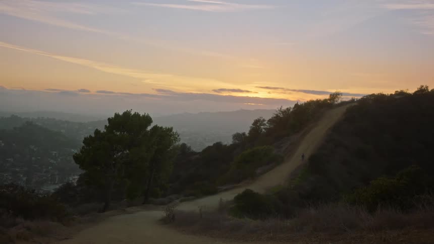 Mountain with a trail and a city in the background with a sunset on the horizon