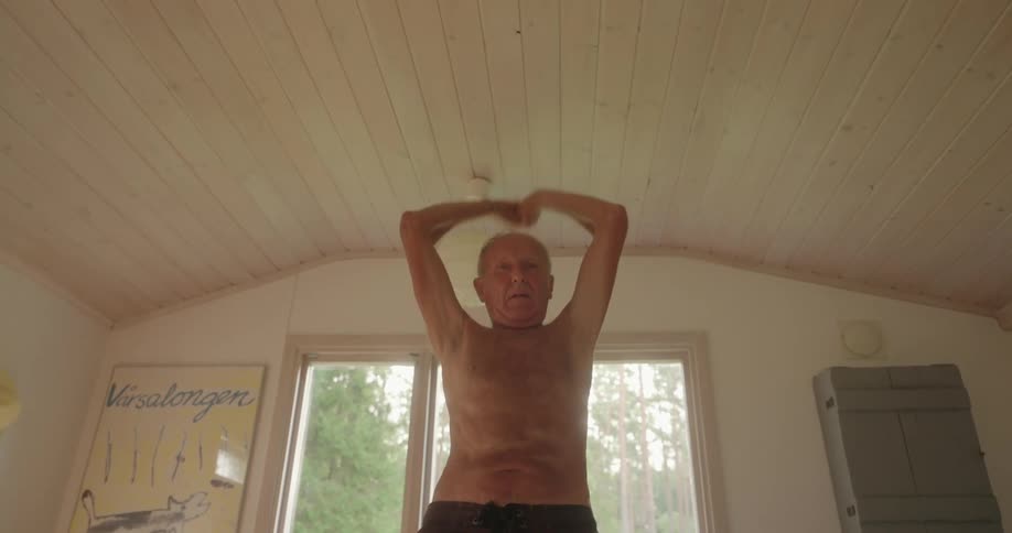 A shirtless elderly man stretches on the living room