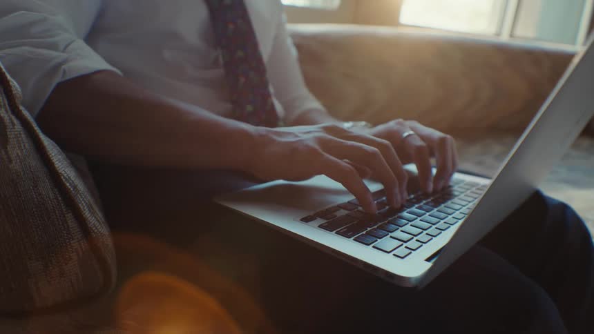 Close up of a mans hands typing on a laptop while sitting on a couch as the sun shines through the window