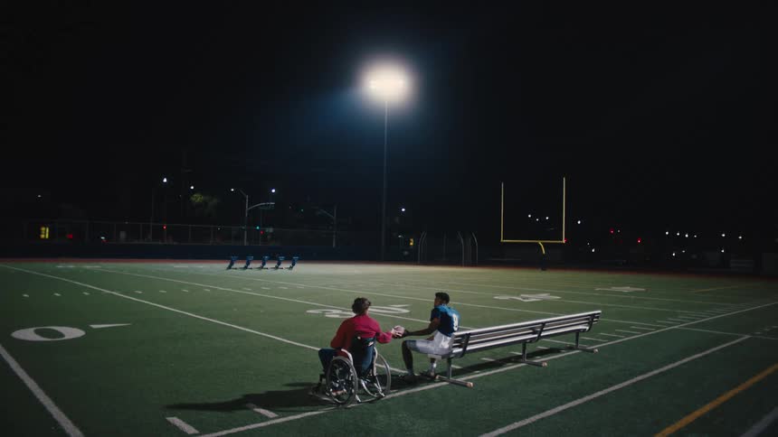 Person in a wheelchair handing the football to a teammate next to him sitting on a bench while both are on a field