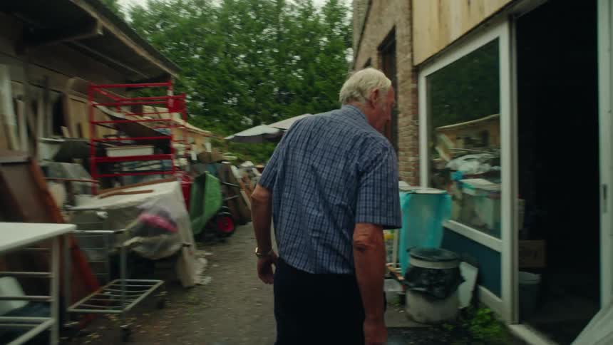Elderly man walking down through a yard surrounded by junk and entering a warehouse with boxes