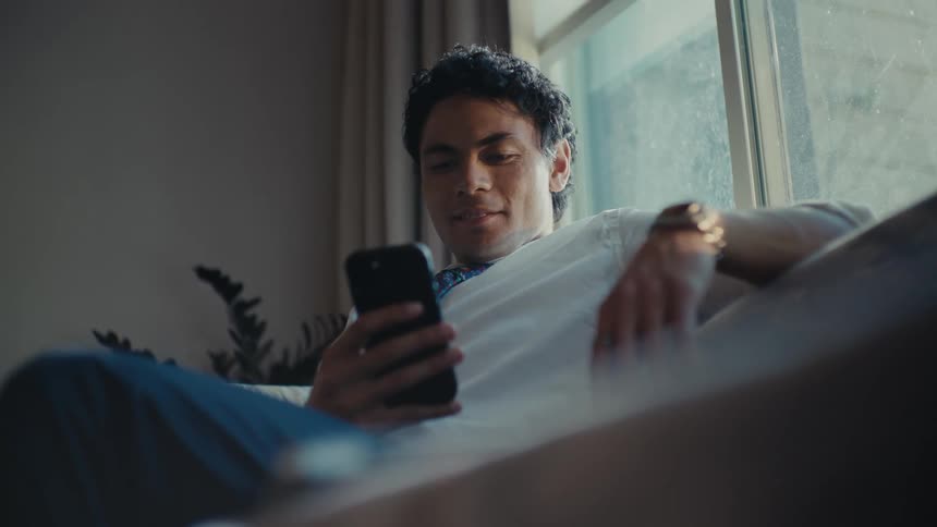 A hispanic man sits on his couch while smiling at his smartphone