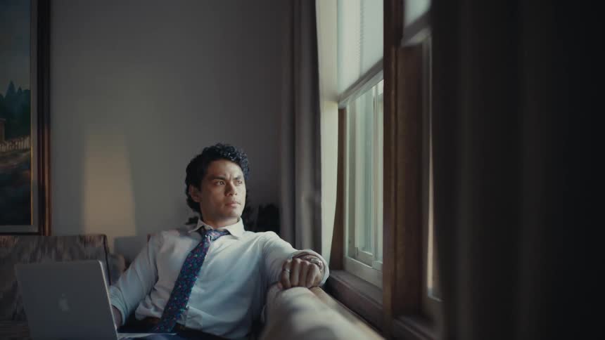 A hispanic man sits on the couch with his laptop while looking out the window