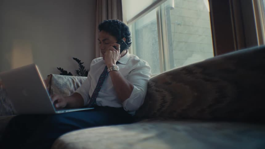 A hispanic man sits on a couch while talking on his smartphone and working on his laptop