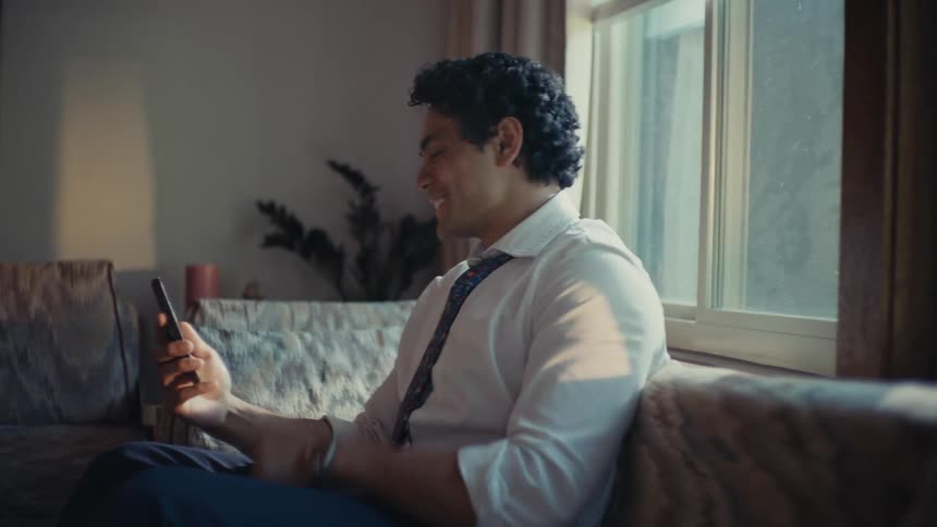 Man in a tie sitting on a sofa making a thumbs up sign to the smartphone in his hands