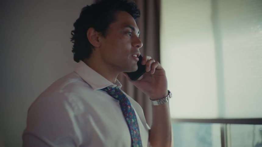 A young man talking on his cellphone held up to his ear while walking around his house holding a glass